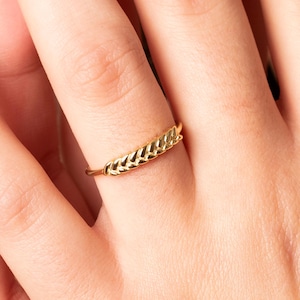 Puede incluir: Un anillo dorado con un diseño de espiga de trigo. El anillo, usado en un dedo, muestra un detallado patrón trenzado. La banda del anillo es delgada, con el diseño de trigo como elemento central. El anillo es de oro.