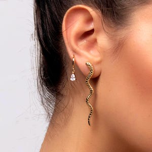 Könnte beinhalten: Goldfarbene Ohrringe. Einer hat einen tropfenförmigen Edelstein an einem kleinen Reifen. Der andere ist ein langes, schlangenartiges Design mit schwarzen Akzenten. Die Ohrringe werden am Ohr einer Person getragen.