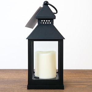 以下が含まれることがあります： ハンドルとクリーム色のろうそくが入った黒い金属製のランタン。ランタンは長方形で、透明なガラスパネルと尖った屋根が付いています。ランタンは木製の表面に置かれています。