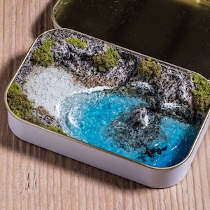 Könnte beinhalten: Eine Miniatur-Landschaftszene in einer weißen Blechdose. Die Szene zeigt einen kleinen blauen See, umgeben von grünem Moos und Steinen.