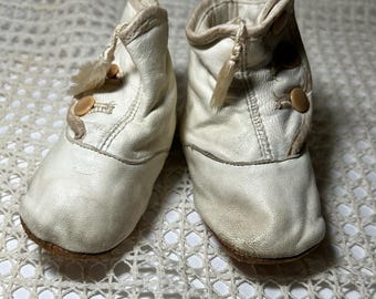 May include: A pair of off-white leather baby booties featuring a high-top design with tan trim, two small round buttons on the side of each shoe, and a decorative tassel detail near the top opening, finished with a soft brown leather sole.
