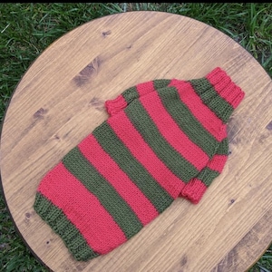 May include: Red and green striped knitted sweater for a pet. The sweater has a turtleneck.