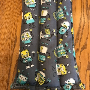 May include: A rectangular, grey fabric heating pad featuring a repeating pattern of colourful cartoon robots. The robots have yellow, blue, and white accents. The pad is vertically divided into sections by stitching.