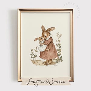 Puede incluir: Una impresión en acuarela enmarcada que presenta dos conejos marrones. Uno de los conejos sostiene al otro, ambos con vestidos. La impresión tiene una estética vintage, con las palabras "Faery Prints" y "Printed & Shipped" visibles.