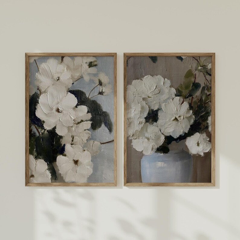 May include: Two framed floral art prints. The paintings feature white flowers with green leaves against a light blue and gray background. One painting shows flowers in a vase. The frames are a light brown color.