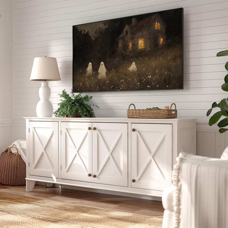 May include: White cabinet with four doors and a woven basket on top. The cabinet has a farmhouse style with X-shaped details on the doors. A painting of a house with ghosts in front of it hangs on the wall above the cabinet.