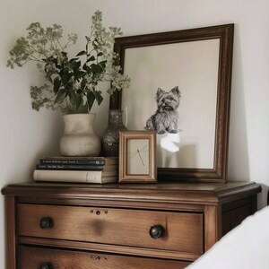 May include: A wooden dresser with three drawers, a framed print of a Yorkshire Terrier, a vase with white flowers, and a small framed print of a dragonfly on top. The dresser is made of dark wood and has a light brown finish.