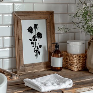 May include: A framed black and white print of a rose bouquet with a wooden frame. The print is on a white background and the roses are in silhouette. The frame is sitting on a wooden countertop with a white towel, a bottle of liquid soap, and a wicker basket.