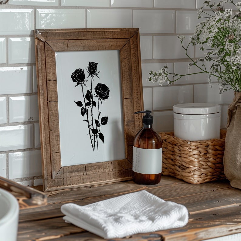 May include: A framed black and white print of a rose bouquet with a wooden frame. The print is on a white background and the roses are in silhouette. The frame is sitting on a wooden countertop with a white towel, a bottle of liquid soap, and a wicker basket.