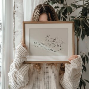 May include: A framed black and white line drawing of a group of sheep lying down in a field. The drawing is in a gold frame and is being held by a person.