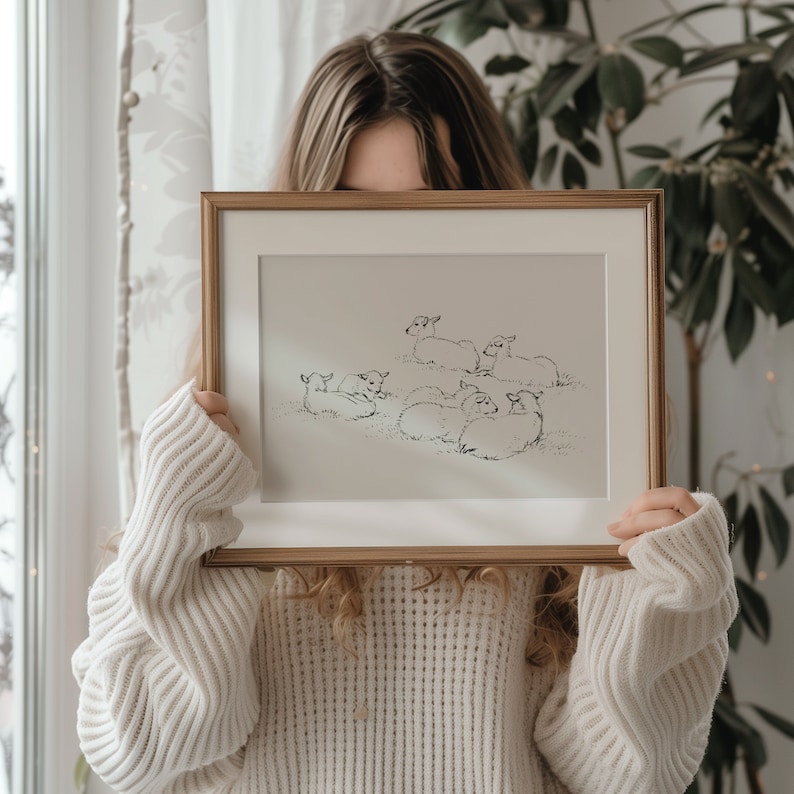 May include: A framed black and white line drawing of a group of sheep lying down in a field. The drawing is in a gold frame and is being held by a person.