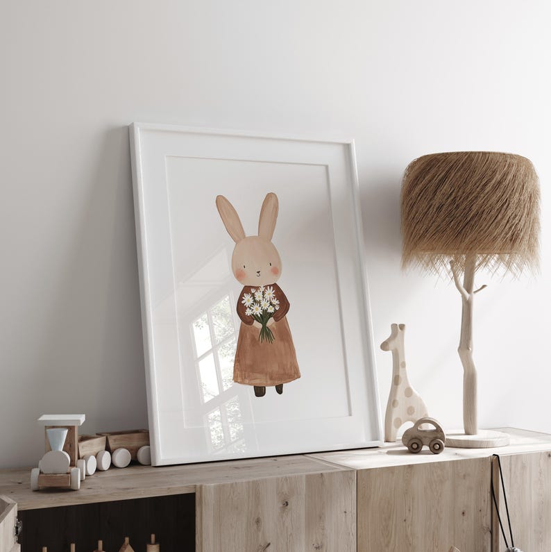 May include: A white framed print of a brown rabbit holding a bouquet of white flowers. The rabbit is wearing a brown dress and is standing on a white background.