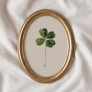 May include: An oval, gold-colored framed print featuring a green four-leaf clover on a beige background. The clover has three visible leaves and a long, thin stem. The frame has a detailed, ornate design.