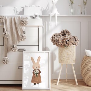 May include: A white framed print of a brown bunny holding a bouquet of white flowers. The bunny is wearing a brown dress. The print is on a white dresser with a brown knitted blanket with white pom-poms.