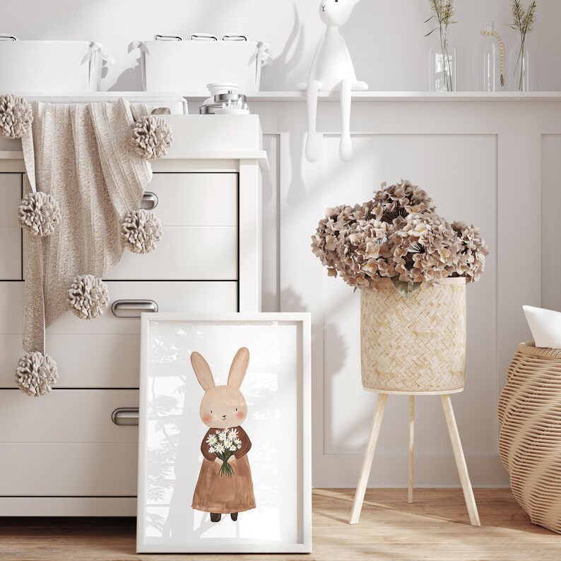 May include: A white framed print of a brown bunny holding a bouquet of white flowers. The bunny is wearing a brown dress. The print is on a white dresser with a brown knitted blanket with white pom-poms.