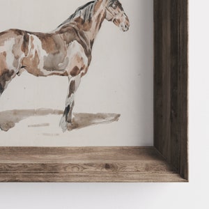 K&ouml;nnte beinhalten: Ein gerahmtes Aquarellbild eines braunen und wei&szlig;en Pferdes, das auf einem wei&szlig;en Hintergrund steht. Der Rahmen ist aus Holz mit brauner Beize.