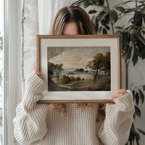 May include: A framed landscape painting with a brown wooden frame. The painting depicts a lake with trees and mountains in the background. The painting is being held by a person.