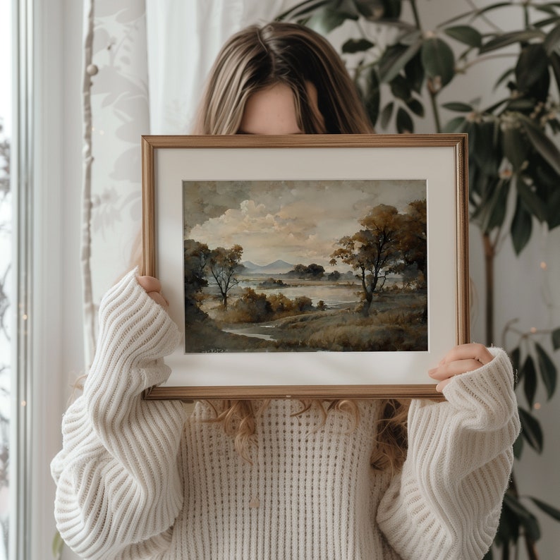May include: A framed landscape painting with a brown wooden frame. The painting depicts a lake with trees and mountains in the background. The painting is being held by a person.