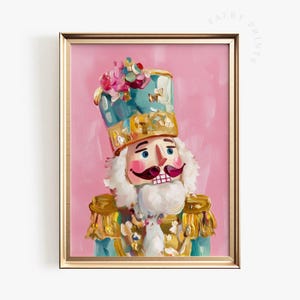 May include: A framed print of a nutcracker soldier with a pink background. The nutcracker has a blue and gold hat, a white beard, and a red mustache. The print is in a gold frame.