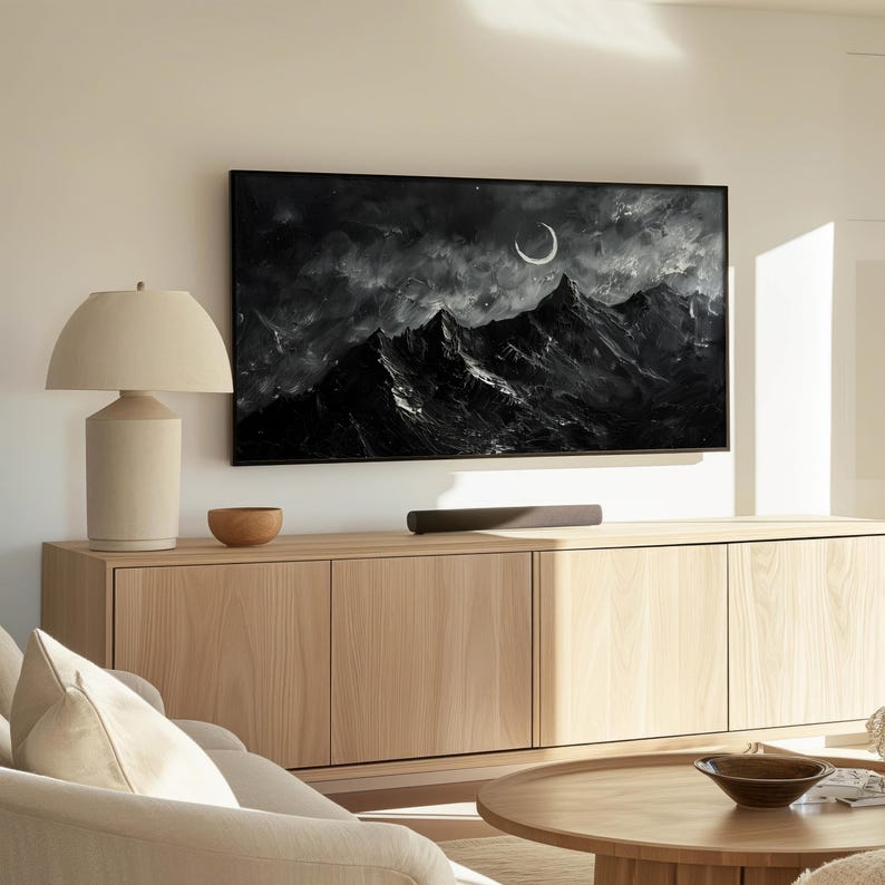 May include: A light wood entertainment center with a flat screen TV displaying a black and white image of a mountain range with a crescent moon. A lamp with a white shade sits on the left side of the entertainment center.
