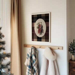 May include: A framed print of Santa Claus in a red coat and white beard, set against a plaid background. Below, a wooden coat rack holds a plaid blanket and a pink coat with a fur-trimmed hood. A Christmas tree is visible on the left.