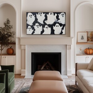 May include: A white fireplace mantel with a black framed painting of white ghosts on a black background. The fireplace has a black hearth and a wood-burning fire. Two tan ottomans are in front of the fireplace. A white couch is on the right side of the image.