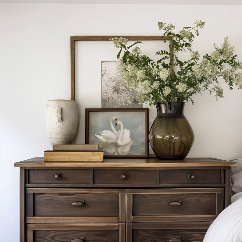 May include: A dark brown wooden dresser with six drawers, a white ceramic vase with a textured surface, a framed print of two white swans, and a bouquet of white flowers in a brown glass vase.