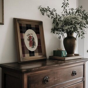 May include: A framed print featuring a vintage illustration of a figure in red and brown attire, set against a plaid background. The wooden frame sits on a dark wood dresser, with a vase of flowers and a wrapped gift nearby.