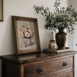 May include: A vintage-style framed painting of pink roses in a teacup, displayed on a dark wood dresser with a vase of white flowers and a small decorative jar.