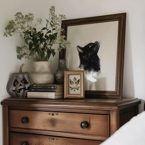 May include: A wooden dresser with three drawers, a black and white cat print, a framed leaf print, a ceramic vase with dried flowers, a pumpkin, and a stack of books.