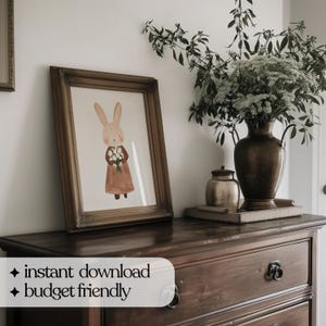 May include: A framed illustration of a brown rabbit holding a bouquet of white flowers. The rabbit is wearing a brown dress and is standing on a light brown background. The frame is a dark brown wood with a gold trim. The image is hanging on a white wall.