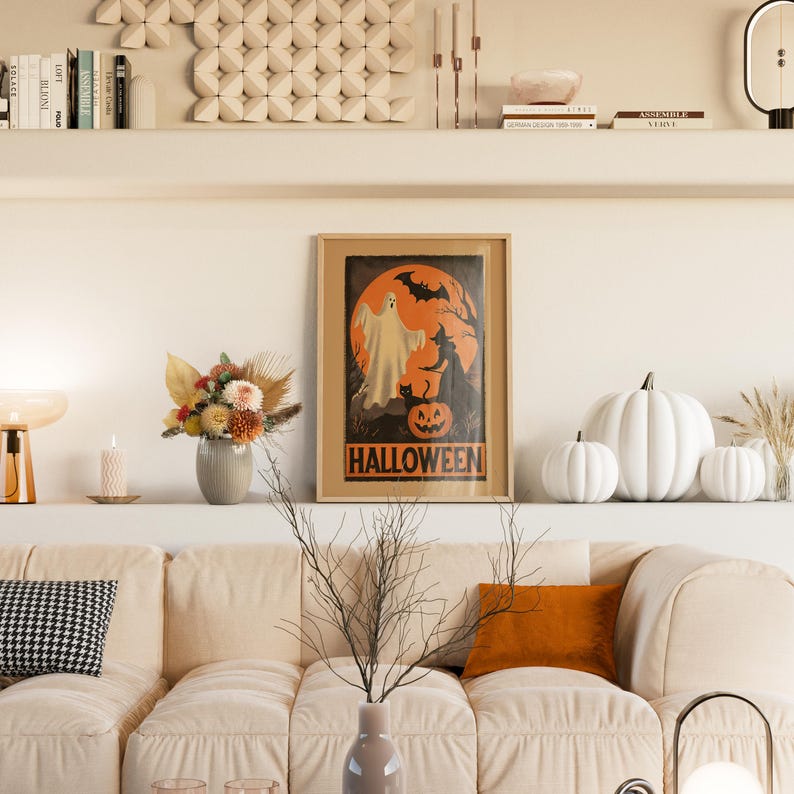 K&ouml;nnte beinhalten: Ein gerahmtes Halloween-Poster mit einem Geist, einer Hexe und einem K&uuml;rbis. Wei&szlig;e K&uuml;rbisse in verschiedenen Gr&ouml;&szlig;en sind auf einem Regal ausgestellt. Im Vordergrund befindet sich ein beiges Sofa mit einem orangefarbenen Kissen und einem schwarz-wei&szlig; gemusterten Kissen.