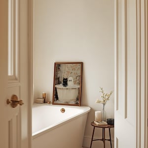 May include: A white bathtub with a gold tap in a bathroom featuring a framed picture of a black cat and a white ghost in a bathtub. A small wooden table with a candle and a vase of white flowers sits next to the bathtub.