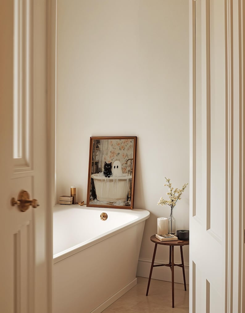 May include: A white bathtub with a gold tap in a bathroom featuring a framed picture of a black cat and a white ghost in a bathtub. A small wooden table with a candle and a vase of white flowers sits next to the bathtub.