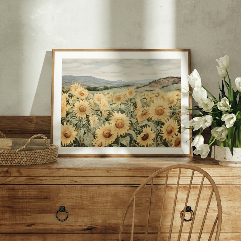 May include: A framed print of a watercolour painting depicting a field of sunflowers in bloom. The sunflowers are yellow with brown centres and green leaves. The background is a soft blue sky with rolling hills in the distance.