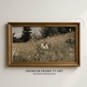 May include: A vintage-style framed print depicting two rabbits in a meadow. The artwork is set in a gold-coloured frame. The scene includes tall grass, wildflowers, and trees. Text at the bottom reads "PREMIUM FRAME TV ART" and "INSTANT DOWNLOAD".