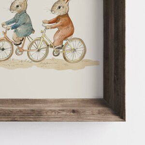 Op de afbeelding: Ingelijste aquarelillustratie van twee konijnen op fietsen. E&eacute;n konijn draagt een blauwe jas, de ander een roodbruin kostuum. De fietsen zijn oranje en geel. De lijst is gemaakt van rustiek bruin hout. Vintage esthetiek.