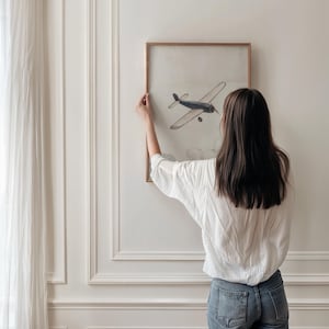 May include: A framed artwork featuring a vintage-style illustration of a blue airplane. The artwork is displayed on a white wall. The frame is a light brown color. The artwork is being hung on the wall.