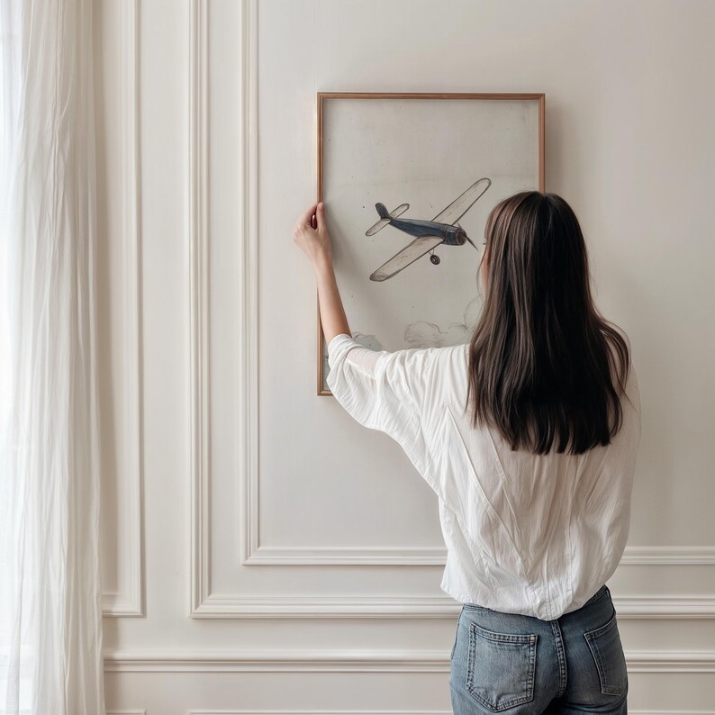 May include: A framed artwork featuring a vintage-style illustration of a blue airplane. The artwork is displayed on a white wall. The frame is a light brown color. The artwork is being hung on the wall.