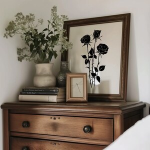 May include: A wooden dresser with three drawers and a black silhouette of roses in a framed print above it. A vase with white flowers sits on top of the dresser, along with a small framed print of a dragonfly.