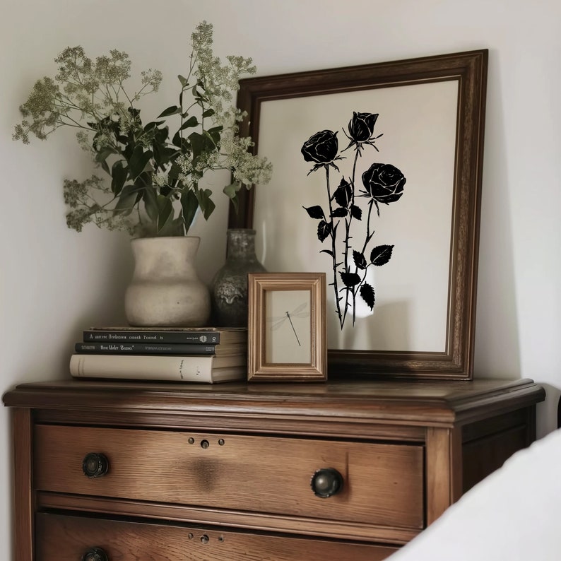 May include: A wooden dresser with three drawers and a black silhouette of roses in a framed print above it. A vase with white flowers sits on top of the dresser, along with a small framed print of a dragonfly.