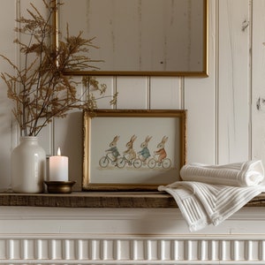Op de afbeelding: Een ingelijste aquarelprint toont vier konijnen op fietsen, tentoongesteld op een rustieke houten plank. Een witte kaars in een messing houder en een opgerolde witte deken staan ook op de plank. Een witte vaas met gedroogde stelen en een goudomrande spiegel maken de sc&egrave;ne compleet.