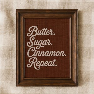 Könnte beinhalten: Ein dunkelbrauner Holzrahmen mit einem tiefroten, strukturierten Hintergrund und dem weißen Text Butter. Sugar. Cinnamon. Repeat. Die Schrift ist in einer kursiven Schreibschrift gehalten und mittig im rechteckigen Rahmen platziert.