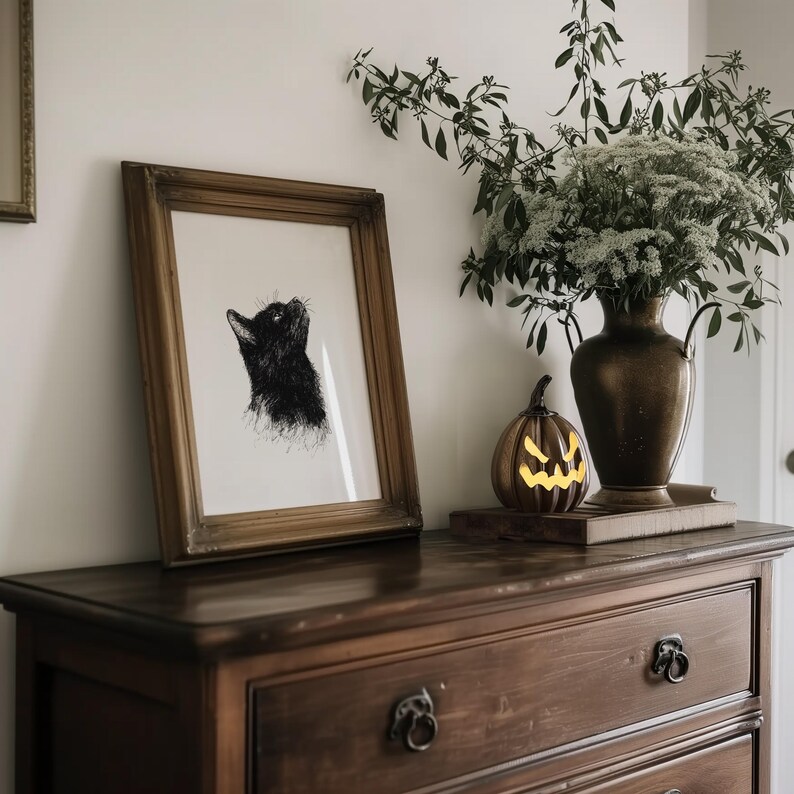 May include: A dark wood dresser with three drawers, a framed black and white illustration of a cat, a small jack-o-lantern, and a vase of white flowers.