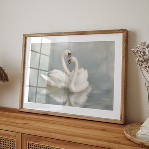 May include: A framed painting of two white swans with their necks intertwined, set against a soft blue and gray background. The swans are reflected in the water below.