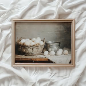 May include: A framed painting of a still life featuring a basket of white eggs and a silver bowl on a wooden table. The background is a muted gray.