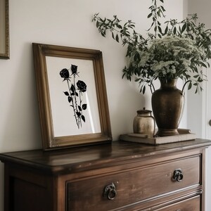May include: A black and white print of three roses in a wooden frame on a dark wood dresser with a vase of white flowers and a small gold jar on the top.