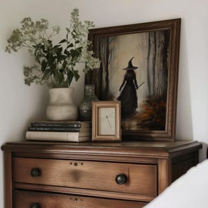 May include: A wooden dresser with three drawers, a framed painting of a witch walking through a forest, a small framed print of a dragonfly, a vase with white flowers, and a stack of books.
