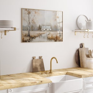 May include: A framed watercolor painting of a white cottage in a field with trees and a stream. The painting has a gold frame and is hanging on a white wall.