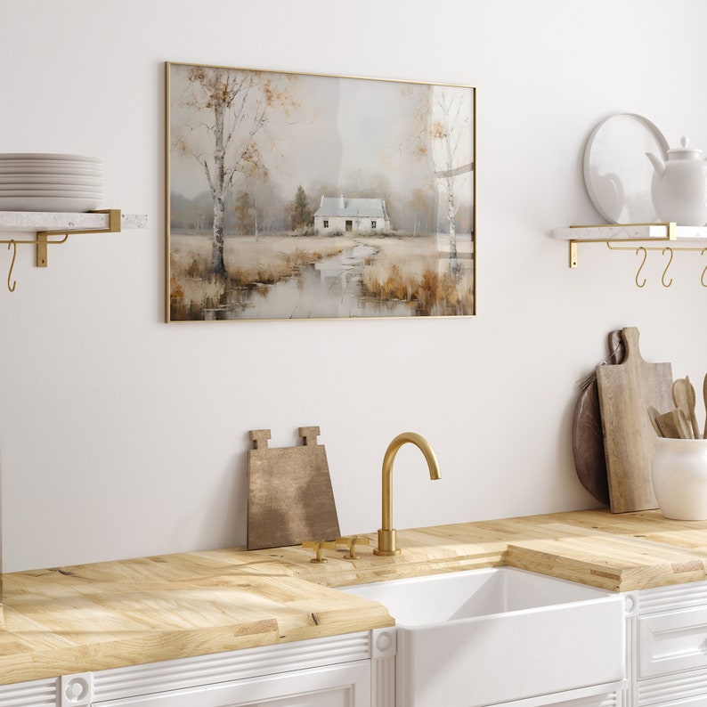 May include: A framed watercolor painting of a white cottage in a field with trees and a stream. The painting has a gold frame and is hanging on a white wall.